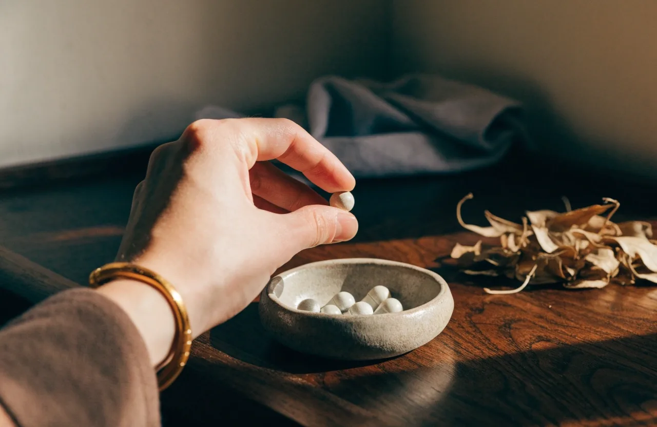 Small ceramic dish of pale capsules on a dark walnut board with a folded cloth and dried botanical leaves